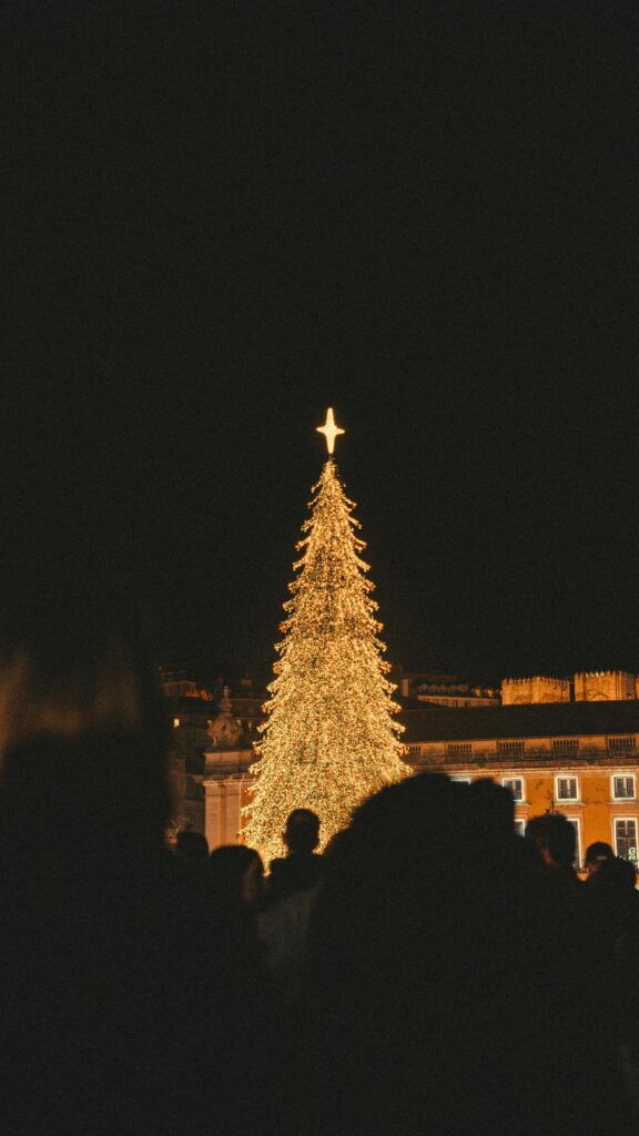 A festive Christmas tree lit up at night in Lisbon, Portugal, surrounded by a crowd.